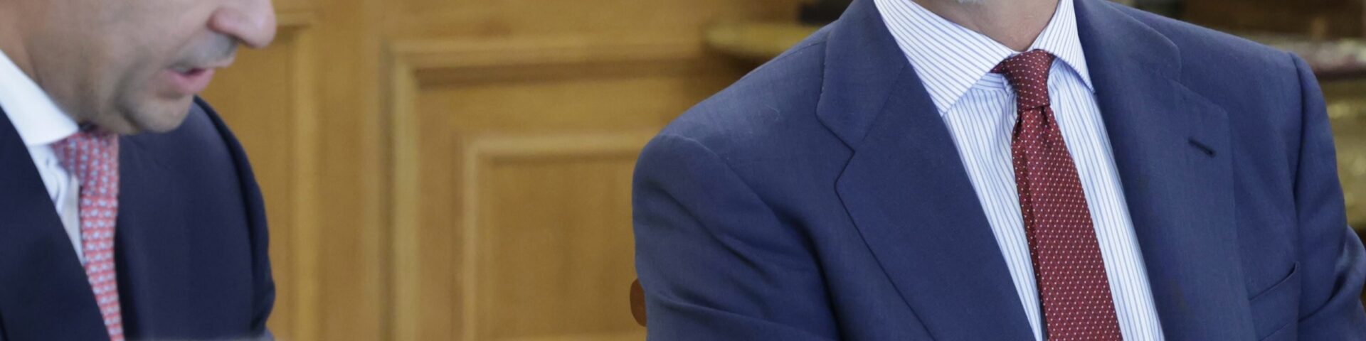 König Felipe VI. sitzend im Palacio de La Zarzuela in Madrid, dunkelblauer Anzug, gestreiftes Hemd und burgunderrote Krawatte mit Punkten, Papiere auf dem Schoß, zuhörend in einem holzgetäfelten Raum - Photo: @casareal.es
