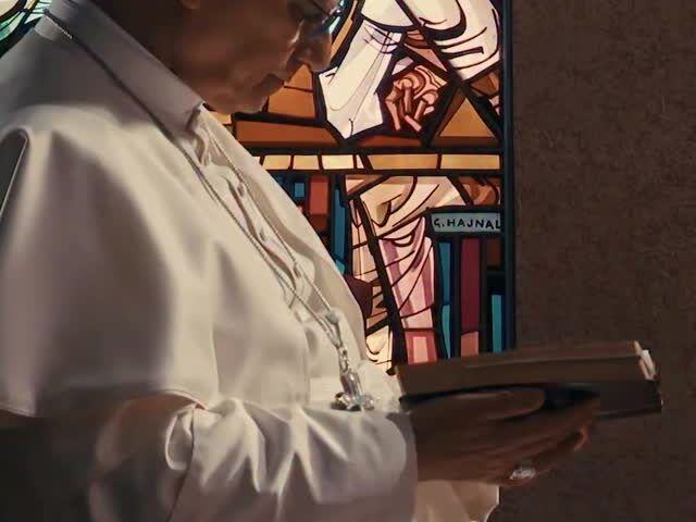 Papst Leo XIV. betet mit gesenktem Haupt und Gebetbuch vor einem bunten Glasfenster.