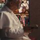 Papst Leo XIV. betet mit gesenktem Haupt und Gebetbuch vor einem bunten Glasfenster.