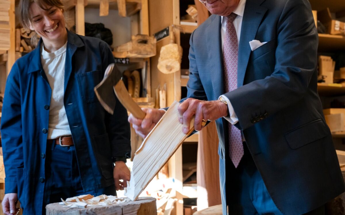 König Carl XVI. Gustaf spaltet im dunklen Anzug Holz auf einem Hackklotz, während Julia Kalthoff in blauer Arbeitsjacke daneben lacht.