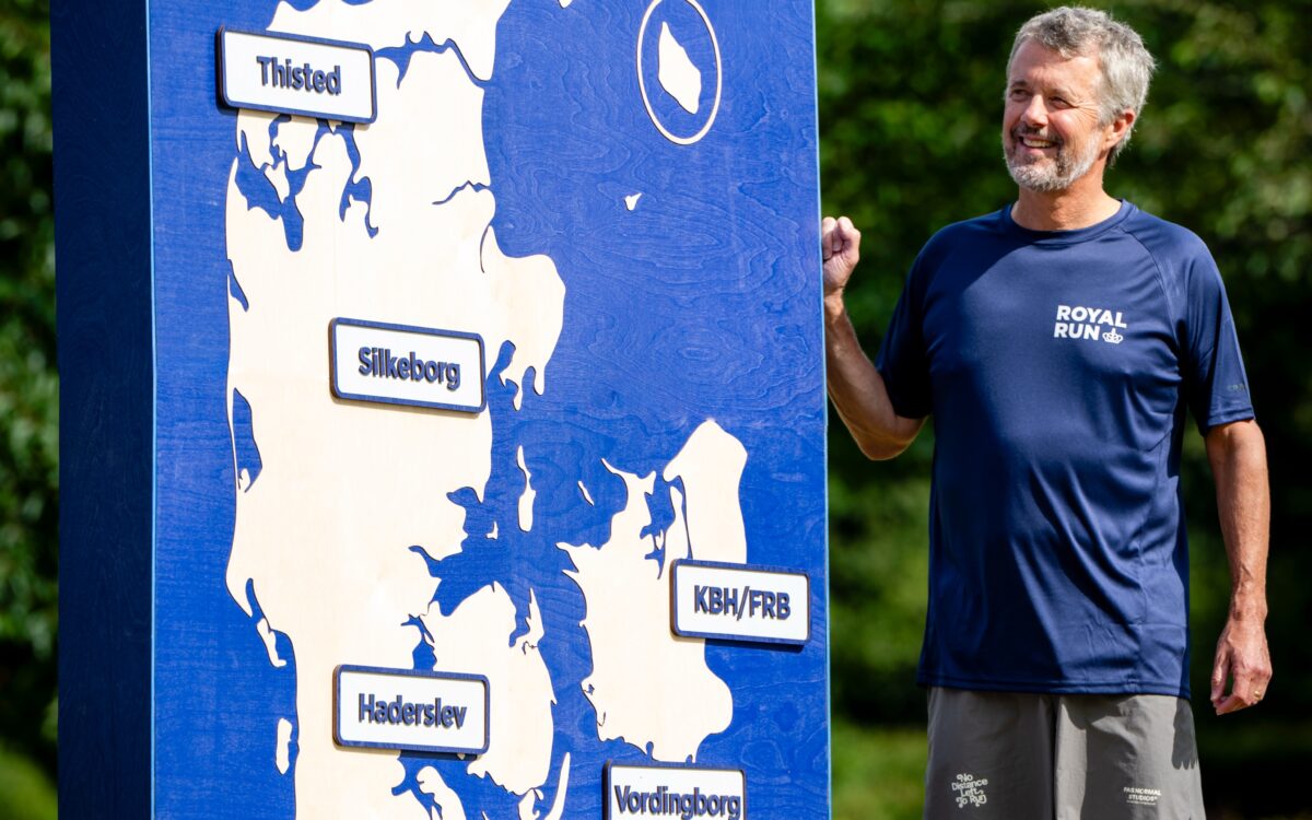 König Frederik X. in navyblauem Royal-Run-Shirt und knallroten Kniestrümpfen neben einer Dänemark-Karte mit den fünf Royal-Run-Städten 2027 — Photo: Matthew James Harrison, Royal Run