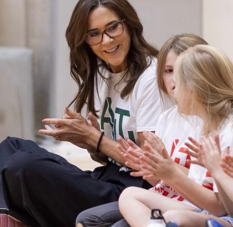Königin Mary in weißem T-Shirt und Brille sitzt lächelnd auf dem Turnhallenboden und klatscht mit Kindern beim Antibulli-Jubiläum — Photo: Mary Fonden