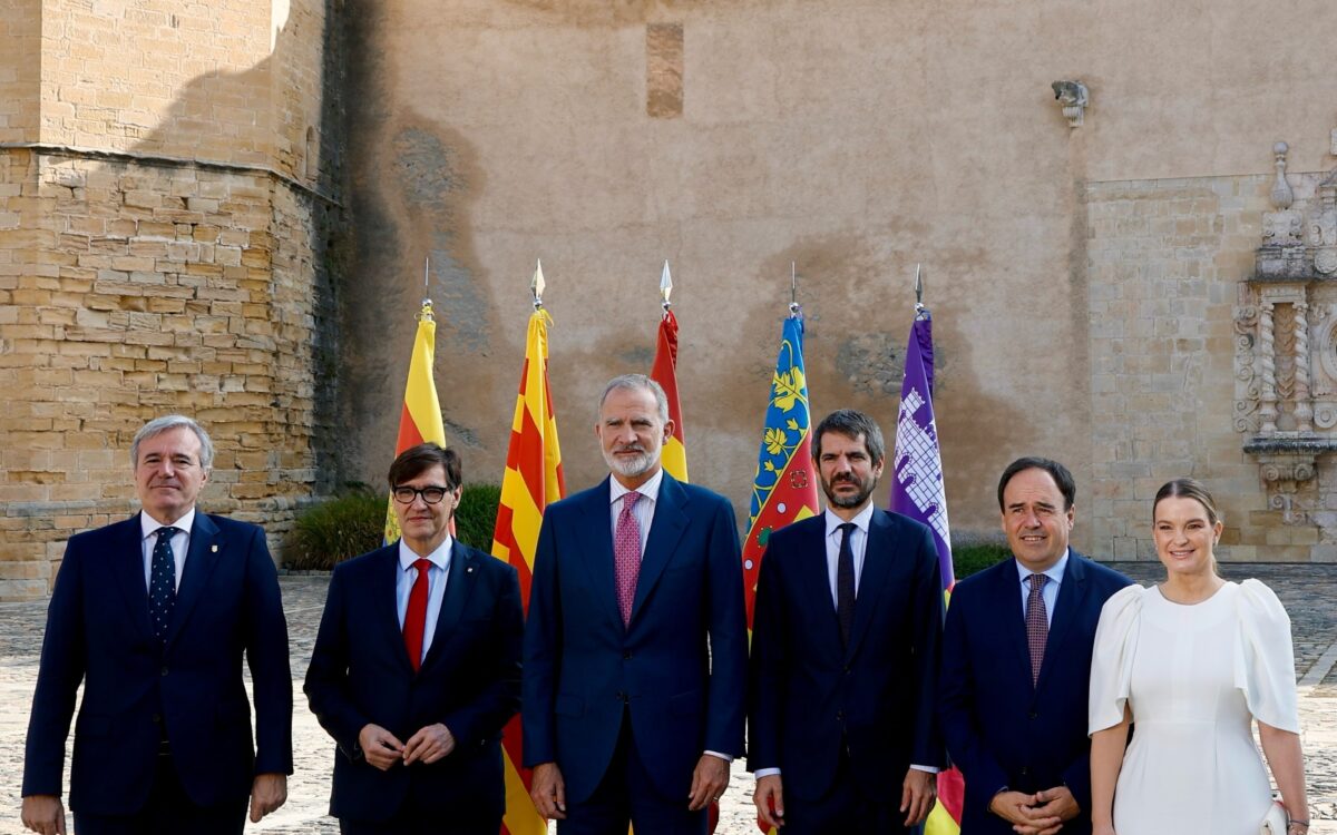 König Felipe VI. in dunkelblauem Anzug mit rosa gemusterter Krawatte steht in der Mitte einer sechsköpfigen Gruppe im gepflasterten Innenhof des Klosters Santa Maria de Poblet, mit Fahnen und honigfarbenen Steinmauern im Hintergrund - Photo: @casareal.es