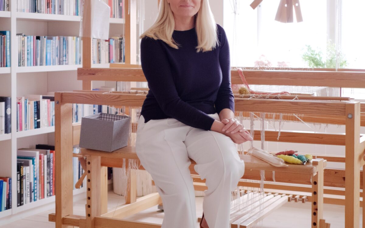Eine Weberin in dunkelblauem Pullover und weisser Hose an einem grossen hölzernen Webstuhl in einem hellen, von Bücherregalen umgebenen Atelier - Foto: Det kongelige hoff