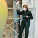 Königin Sonja glasiert in dunkelgrünem Arbeitsanzug und mit Maske auf einem Gerüst einen riesigen gelb-orangen Keramiktopf - Foto: Det kongelige hoff