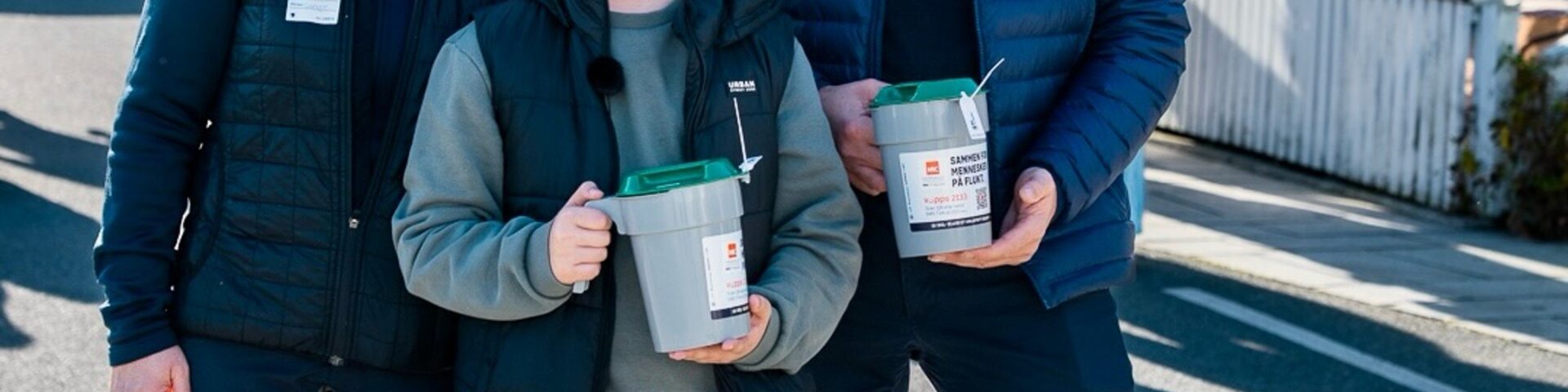 King Haakon smiling on a sunny street on Kvitsoy holding a TV-aksjonen collection bucket beside a young volunteer and Camilla - Photo: Det kongelige hoff
