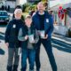 King Haakon smiling on a sunny street on Kvitsoy holding a TV-aksjonen collection bucket beside a young volunteer and Camilla - Photo: Det kongelige hoff