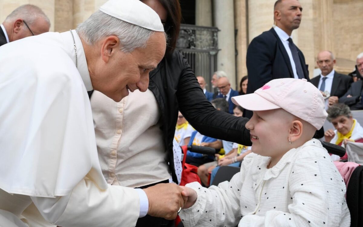 Papst Leo XIV. beugt sich auf dem Petersplatz lächelnd zu einem Mädchen mit rosa Kappe hinab und hält ihre Hand.