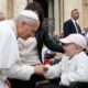 Papst Leo XIV. beugt sich auf dem Petersplatz lächelnd zu einem Mädchen mit rosa Kappe hinab und hält ihre Hand.