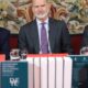 König Felipe VI. in der Mitte eines Tisches im Königspalast La Granja de San Ildefonso in Segovia, dunkler Nadelstreifenanzug und geblümte Krawatte, vor ihm die Bände des Diccionario Historico de la Lengua Espanola und hinter ihm ein historischer Wandteppich - Photo: @casareal.es