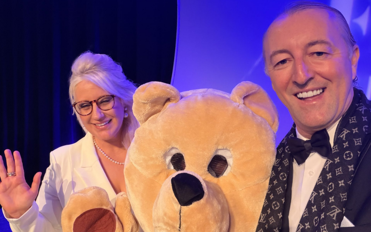 Prinz Mario-Max zu Schaumburg-Lippe laechelt mit einer Mitstreiterin und einem grossen Teddybaer-Maskottchen bei einer Wohltätigkeitsveranstaltung in blauem Buehnenlicht.