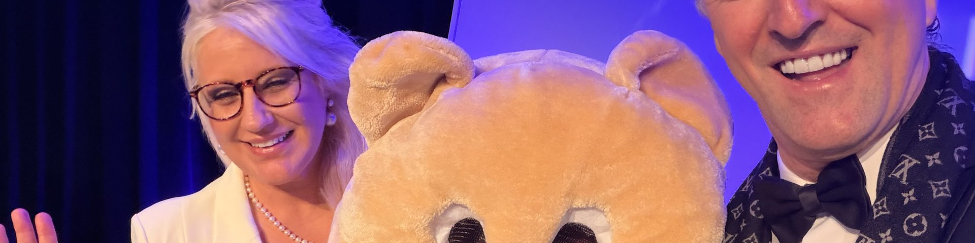 Prinz Mario-Max zu Schaumburg-Lippe laechelt mit einer Mitstreiterin und einem grossen Teddybaer-Maskottchen bei einer Wohltätigkeitsveranstaltung in blauem Buehnenlicht.