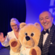 Prinz Mario-Max zu Schaumburg-Lippe laechelt mit einer Mitstreiterin und einem grossen Teddybaer-Maskottchen bei einer Wohltätigkeitsveranstaltung in blauem Buehnenlicht.