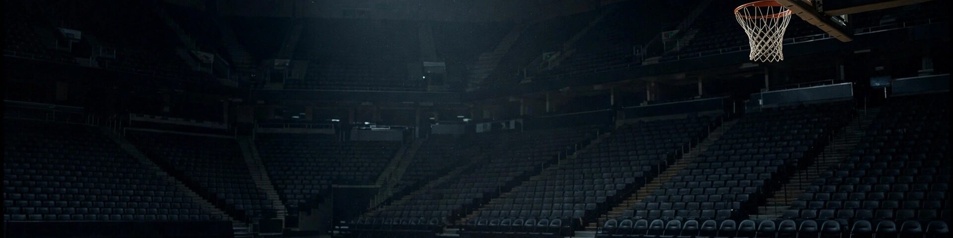 Basketball-Arena bei Nacht, ein Spotlight auf leerem Court, ein Basketball fliegt auf den Korb zu.