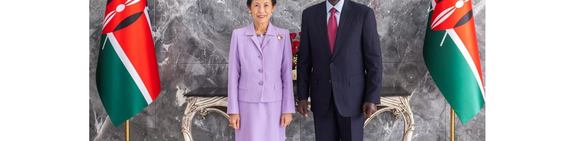 Prinzessin Hisako von Takamado in einem fliederfarbenen Kostuem neben Kenias Praesident William Ruto zwischen zwei kenianischen Flaggen unter dem Wappen Kenias in Nairobi - Foto: @kunaicho_jp